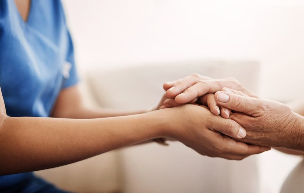 Close-up of a compassionate caregiver in blue scrubs holding an elderly person's hands to offer comfort and support."